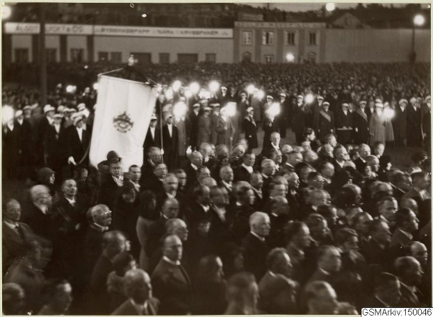 Schwarzes und weißes Foto einer großen Menge vor einem Gebäude, mit einer Person, die eine Fahne hält, in der Mitte, Polen und Lichter im Hintergrund und Text, der unten 'Gustav Klimts Rede an der Berliner Mauer' lautet.