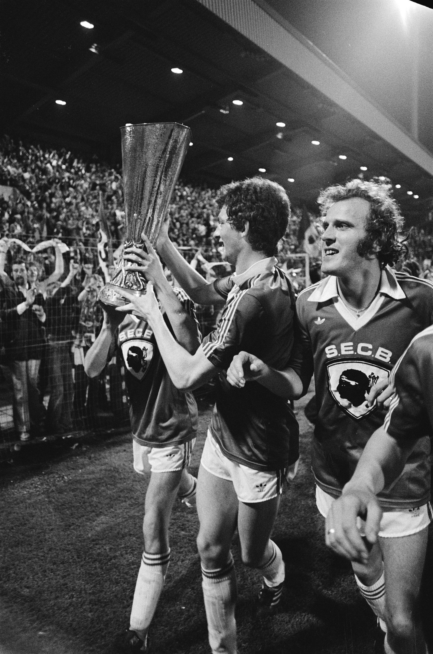 Schwarzes und weißes Foto von Fußballspielern, die mit einem Pokal auf einem Feld feiern, umgeben von einer Menge und Stadionbeleuchtung.