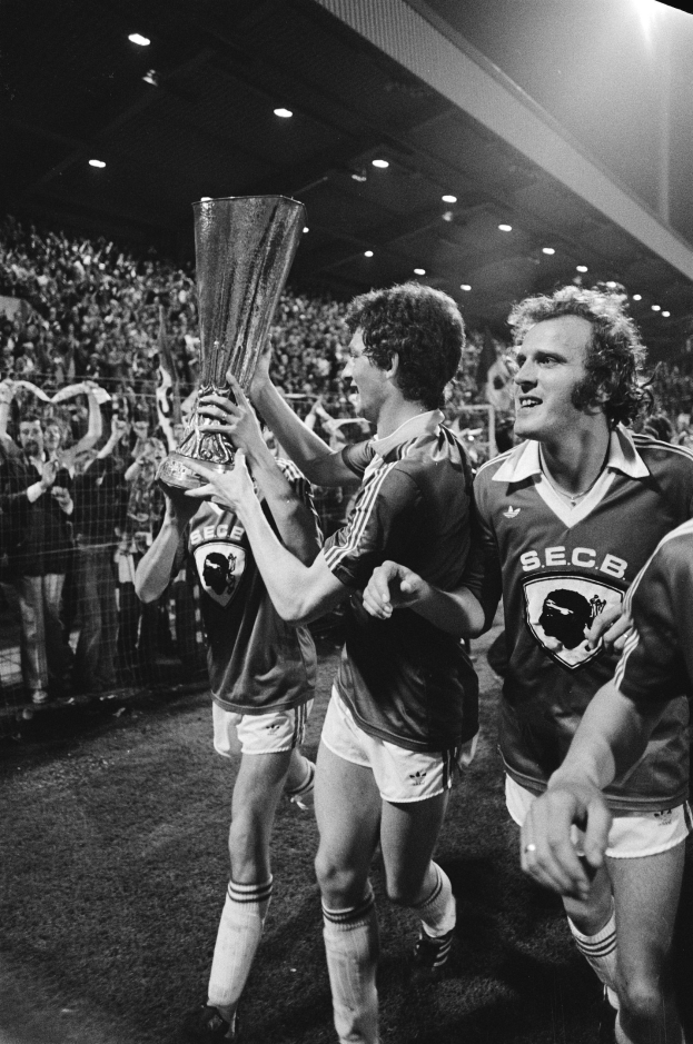 Schwarzes und weißes Foto von Fußballspielern, die mit einem Pokal auf einem Feld feiern, umgeben von einer Menge und Stadionbeleuchtung.