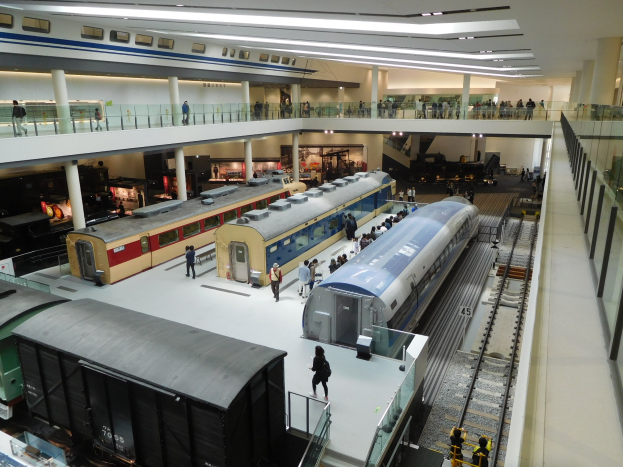 Ein großer Museumsraum mit verschiedenen Zügen in unterschiedlichen Gröüen und Farben, mit Menschen, die die Ausstellungsstücke und architektonische Elemente wie Geländer, Pfeiler und Deckenleuchten betrachten.