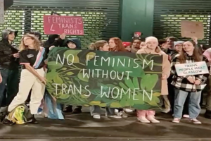 Eine Gruppe von Menschen hält ein Banner mit der Aufschrift "Kein Feminismus ohne Trans-Frauen" und verschiedene Schilder in einem öffentlichen Raum mit einer Wand und einem Rollladen im Hintergrund.