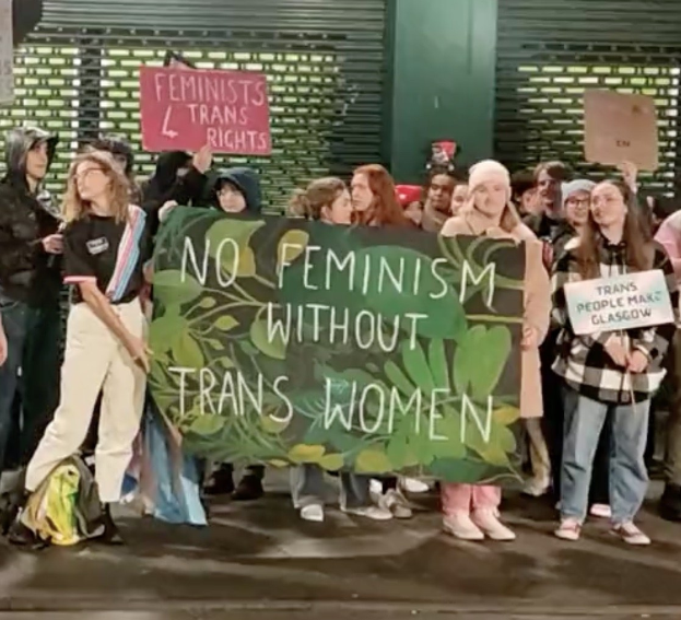 Eine Gruppe von Menschen hält ein Banner mit der Aufschrift "Kein Feminismus ohne Trans-Frauen" und verschiedene Schilder in einem öffentlichen Raum mit einer Wand und einem Rollladen im Hintergrund.