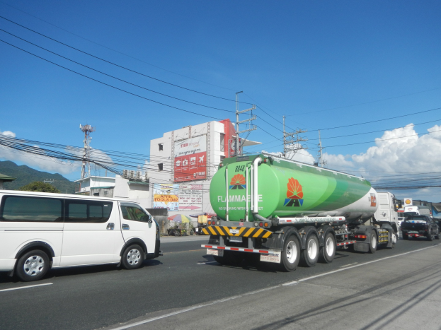 Ein großer Tanklastzug fährt auf einer Straße neben einem weißen Van, mit Strommasten, Gebäuden, Werbetafeln, Bäumen, Hügeln und einem bewölkten Himmel im Hintergrund.