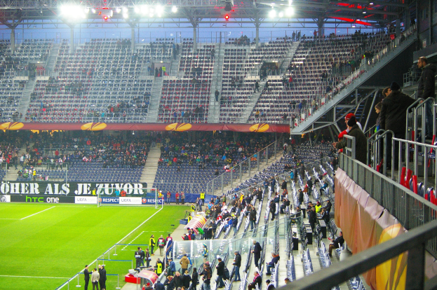 Großes Stadion voller Zuschauer bei einem Fußballspiel, mit sitzenden und stehenden Personen, einem Geländer, einer Fahne und einem leeren Abschnitt im Hintergrund mit Deckenbeleuchtung.