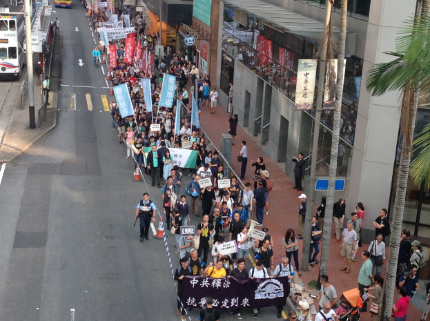 Eine große Gruppe von Menschen marschiert auf einer Straße in Hong Kong, mit Protestschildern in den Händen, mit Bäumen, glaswandigen Gebäuden und Fahrzeugen auf der Straße.