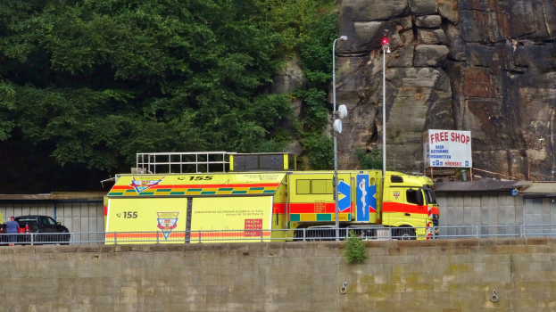 Ambulanz am Rande einer Straße neben einer Klippe geparkt, mit verschiedenen Objekten im Vordergrund und Bäumen und einem Auto im Hintergrund.