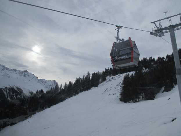 Eine Seilbahn fährt einen schneebedeckten Berg hinauf, mit Bäumen im Hintergrund, unter einem bewölkten Himmel mit hellerer Sonne.