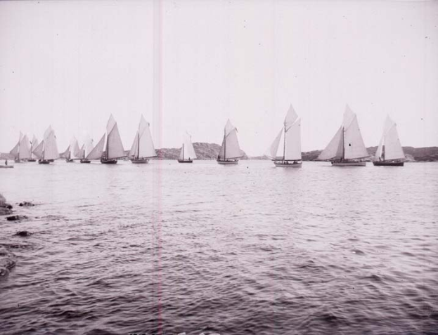 Ein Schwarz-Weiß-Foto mehrerer Segelboote auf dem Wasser mit Hügeln im Hintergrund und einem klaren Himmel, mit Text unten, das es als Foto des New York Yacht Club identifiziert.