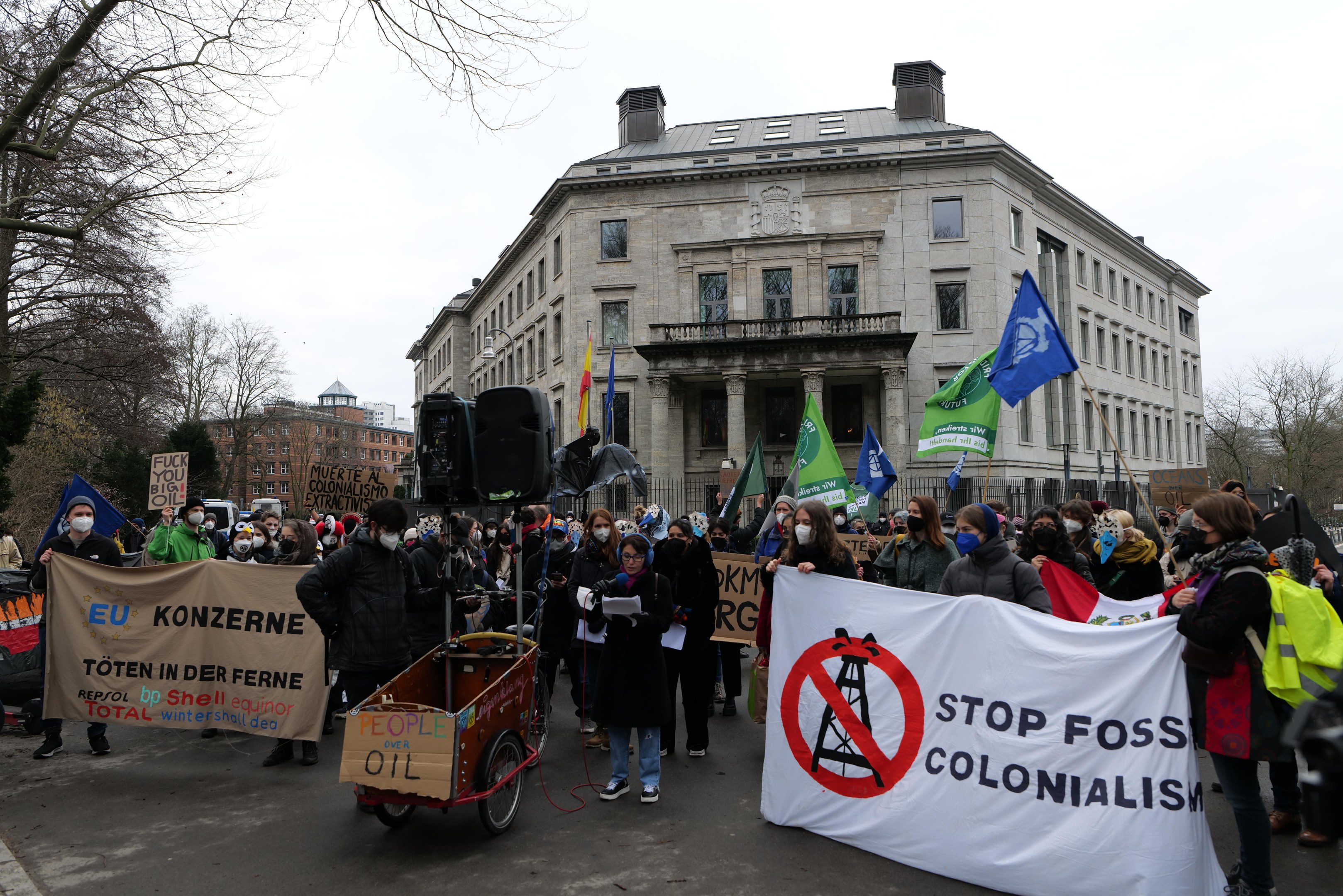 Große Gruppe von Menschen marschiert auf einer Straße bei einer Protestaktion gegen fossile Brennstoffe, hält Schilder und Fahnen, mit einem Fahrzeug im Vordergrund und Gebäuden, Bäumen und einem klaren blauen Himmel im Hintergrund.