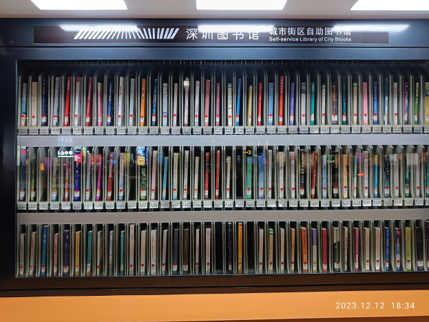 Eine gut beleuchtete Bibliothek mit hohen Bücherregalen voller Bücher, die ein Schild mit Text und ein Wasserzeichen am unteren Rand zeigt, das an eine Selbstbedienungsbibliothek in einer städtischen Umgebung erinnert.