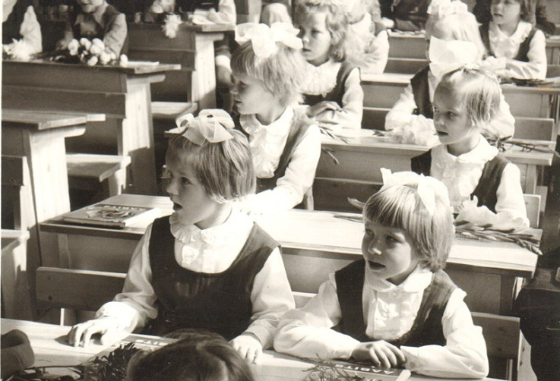 Schwarzes und weißes Foto von jungen Mädchen in Schuluniformen, die an aufgeräumt angeordneten Tischen in einem Klassenzimmer sitzen.