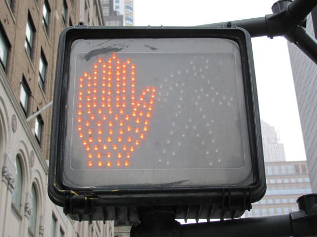 Eine Ampel mit rotem Handzeichen in einer städtischen Umgebung mit Gebäuden auf beiden Seiten und einem klaren Himmel darüber.