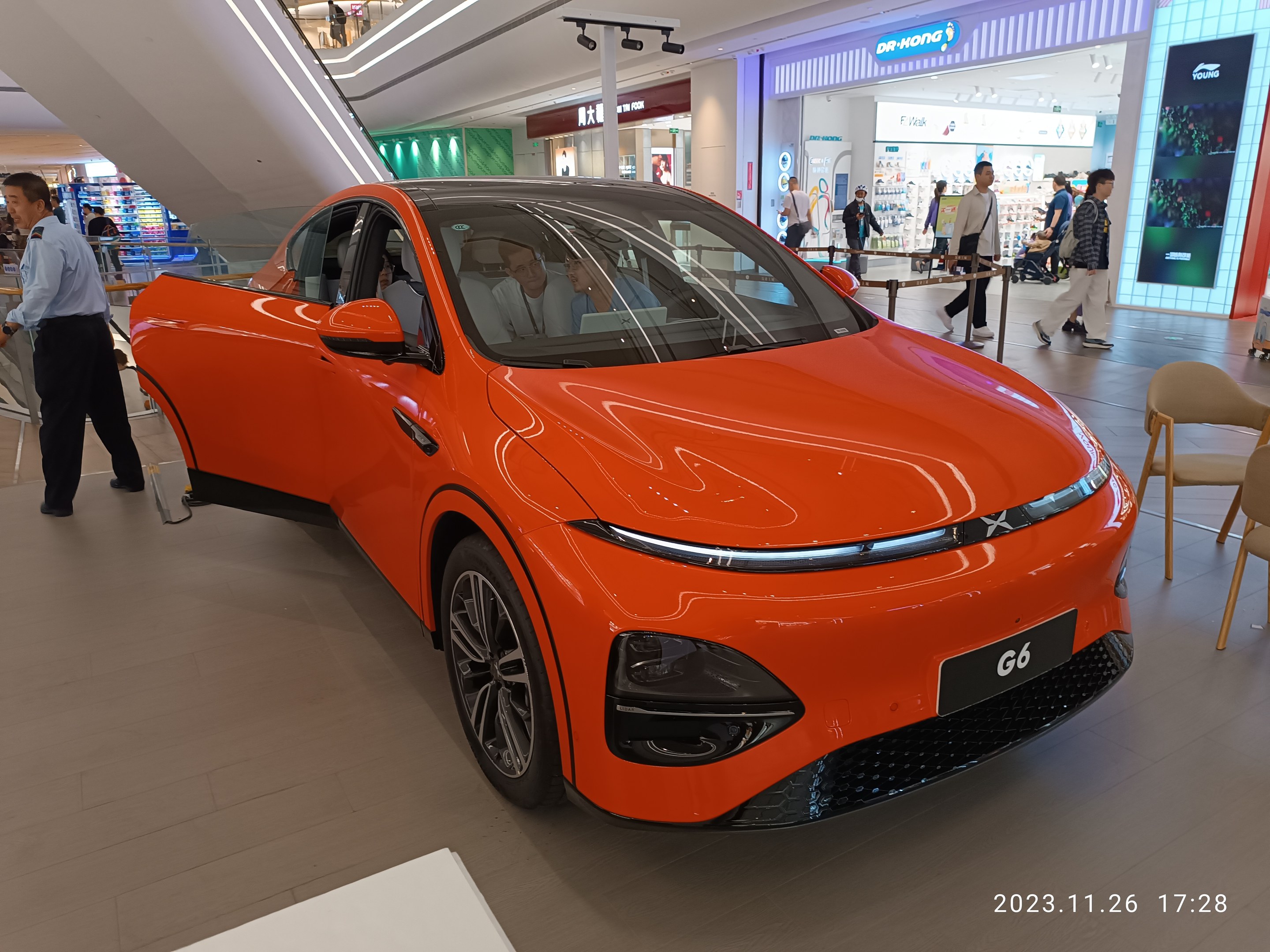 Oranges Elektroauto in einer Ausstellungshalle umgeben von Menschen, Stühlen, einem Wagen und Ladenschildern im Hintergrund.