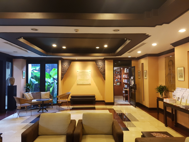Ein geräumiger Hotelfoyer mit Sitzbereichen, Dekorationspflanzen, Bücherregalen, Leuchten, Wandgemälden und Beistelltisch-Dekoration, beleuchtet von natürlichem Licht durch Fenster und Türen.