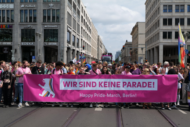 Eine Gruppe von Menschen marschiert auf einer Straße in Berlin, Deutschland, mit einer pinken 'Happy Pride March'-Fahne, während Gebäude, Laternenpfähle und Verkehrszeichen die Straße säumen, unter einem bewölkten Himmel.