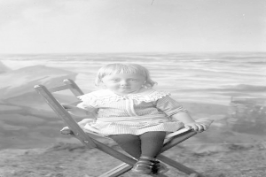 Ein kleines Mädchen sitzt in einem Stuhl am Strand, mit Meer und Himmel im Hintergrund, auf einem alten Schwarz-Weiß-Foto.
