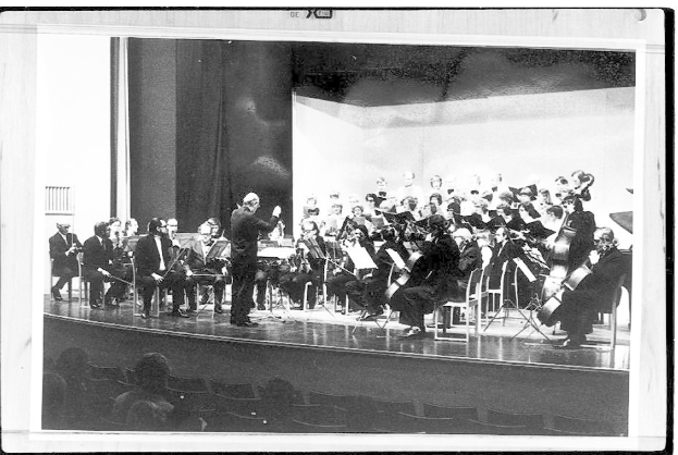 Ein Schwarz-Weiß-Foto eines Dirigenten in Anzug und Krawatte auf einer Bühne, der ein Orchester mit Geigen, Celli und Flöten leitet, während das Publikum auf Stühlen aufmerksam zuhört.