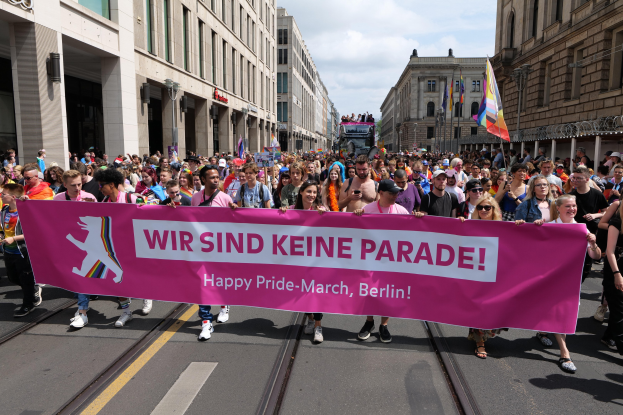 Gruppe von Menschen marschiert auf einer Berliner Straße mit einem pinken 'Happy Pride March'-Schild, mit Gebäuden an der Straße und weißen Wolken am Himmel.