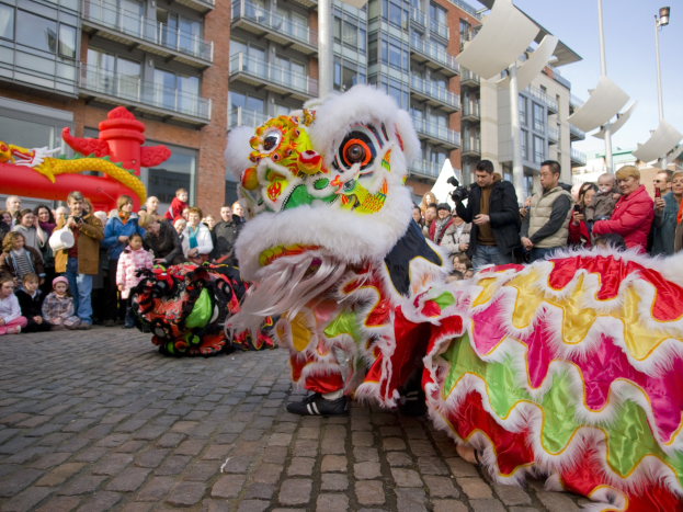 Ein lebendiges chinesisches Neujahrsfest in Amsterdam mit einem Löwen tanzen im Vordergrund und einer Menge Menschen, einige halten Kameras, drum herum und Gebäuden, Laternenmasten und einem klaren blauen Himmel im Hintergrund.