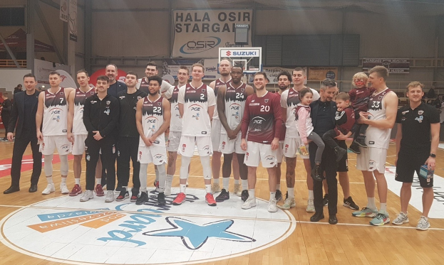 Gruppe von Männern und Kindern auf einem Basketballfeld mit Schildern "Hala Osir Starga", sitzende Zuschauer, ein Basketballkorb, Metallstangen, Deckenleuchten und eine Wand im Hintergrund.