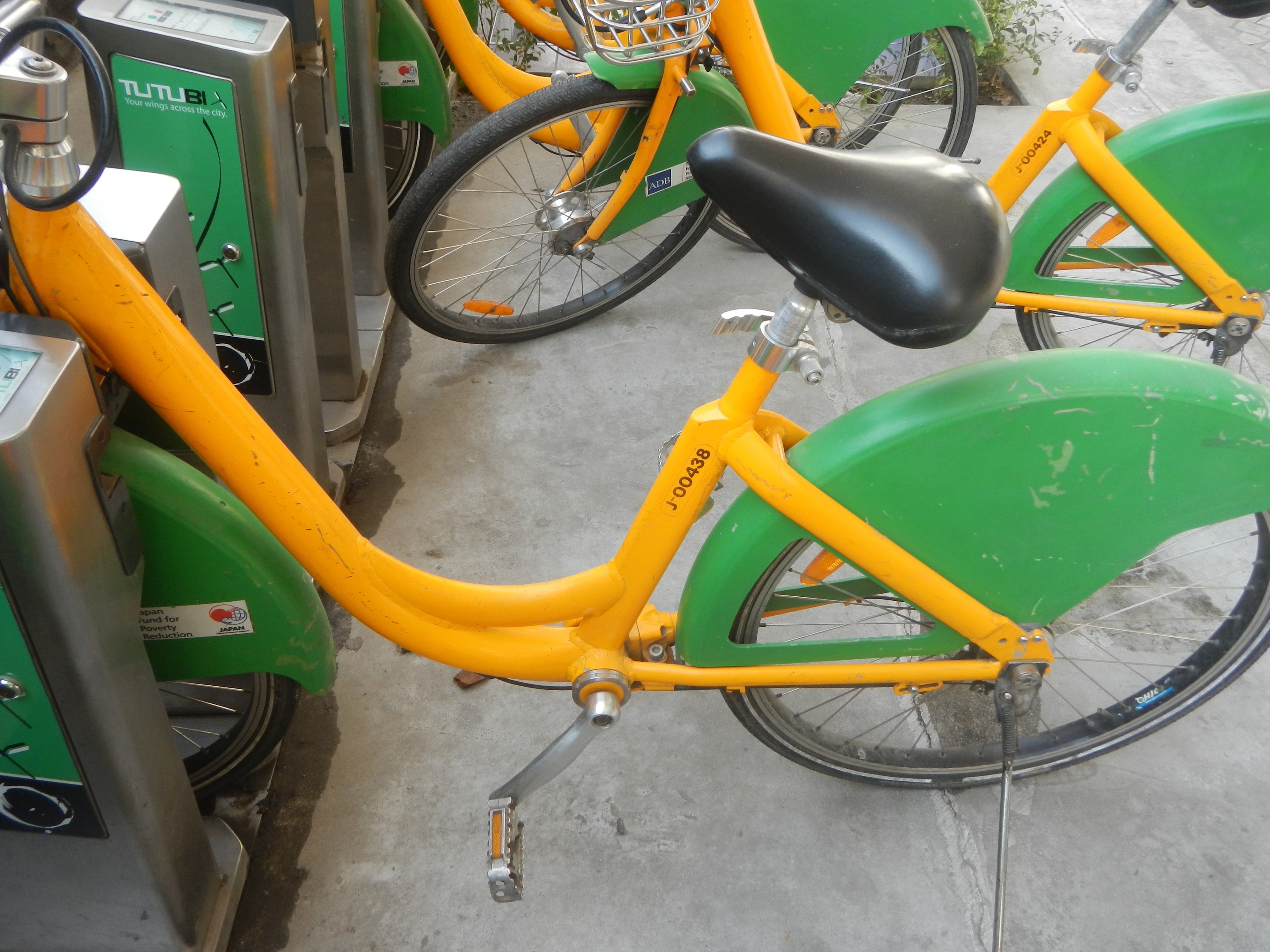 Eine Reihe gelber und grüner Fahrräder nebeneinander auf dem Boden geparkt, mit Maschinen und Pflanzen im Hintergrund.