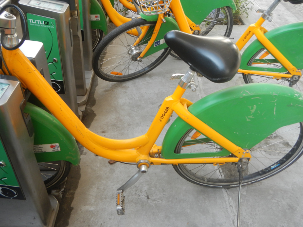 Eine Reihe gelber und grüner Fahrräder nebeneinander auf dem Boden geparkt, mit Maschinen und Pflanzen im Hintergrund.