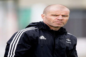 Ein kahlköpfiger Trainer in einer schwarzen Jacke steht auf einem Fußballfeld, trägt eine marineblaue Jacke mit weißen Streifen und einem weißen Kragen und hat die Hände gefaltet und einen ernsten Gesichtsausdruck.