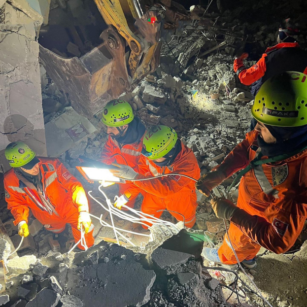 Gruppe von Menschen in orangefarbenen Uniformen und grünen Helmen bei der Arbeit an Schutt von einem eingestürzten Gebäude, mit Werkzeugen und Materialien, mit einem Kran im Hintergrund.