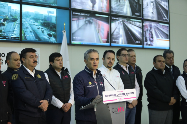 Gruppe von Männern vor einem Rednerpult mit einem Mikrofon und einer Tafel mit der Aufschrift "Mexikanische Zoll- und Grenzschutzbehörde", mit Fernsehern und einer Flagge an der Wand dahinter.