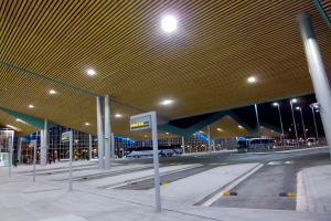 Eine Bushaltestelle nachts mit hellen Deckenleuchten, Fahrzeugen auf der Straße, Beschilderung, einigen Menschen und beleuchteten Gebäuden im Hintergrund.