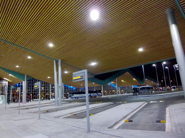 Eine Bushaltestelle nachts mit hellen Deckenleuchten, Fahrzeugen auf der Straße, Beschilderung, einigen Menschen und beleuchteten Gebäuden im Hintergrund.