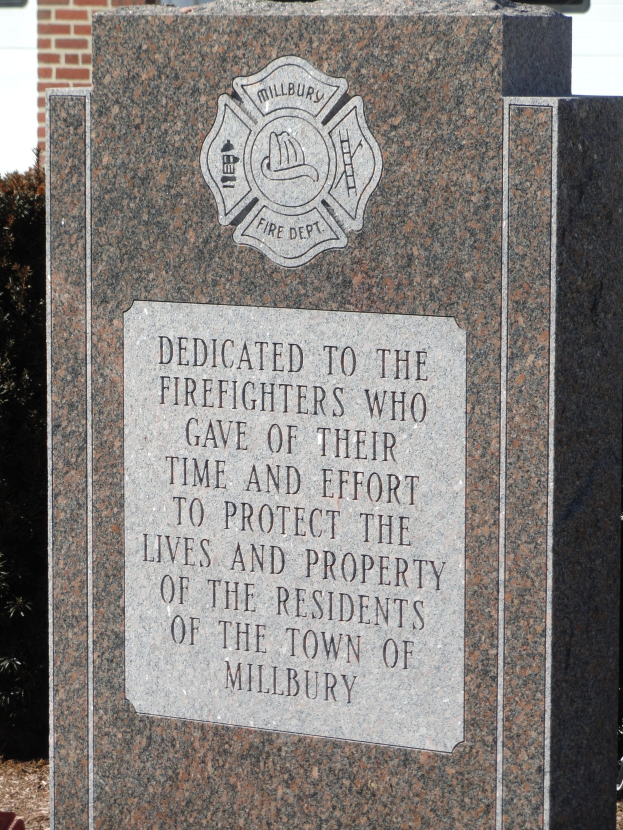 Denkmalstein mit einer Gedenktafel für Feuerwehrleute in Millbury, vor einem Backsteingebäude mit umliegender Vegetation.