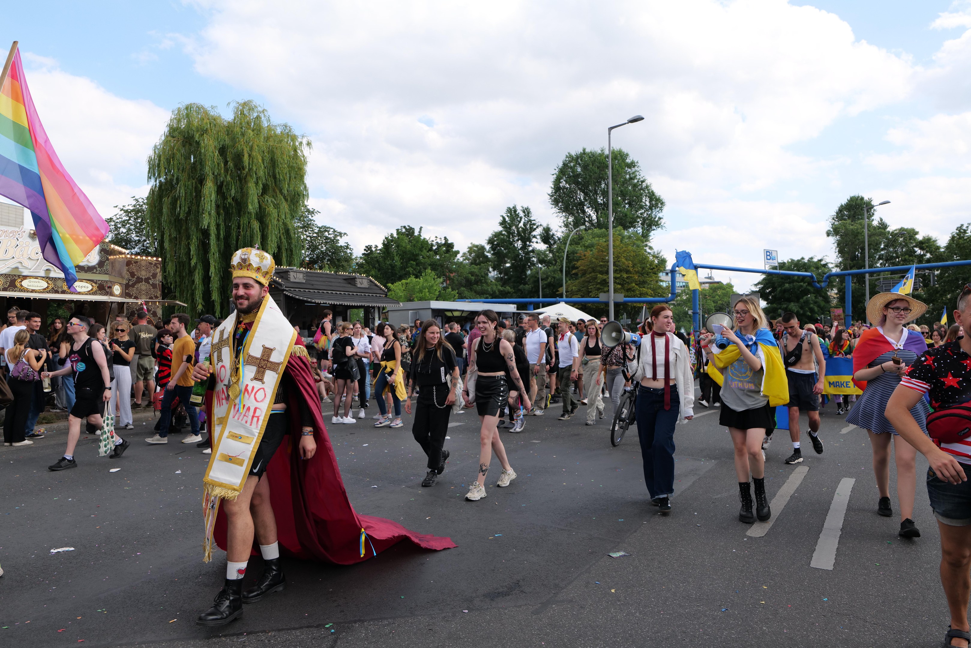 Eine Gruppe von Menschen marschiert bei der Gay Pride Parade 2018 mit einer Regenbogenflagge und Musikinstrumenten, einige tragen Mützen, vorbei an Laternenmasten, Bäumen, Schuppen und einem bewölkten Himmel.