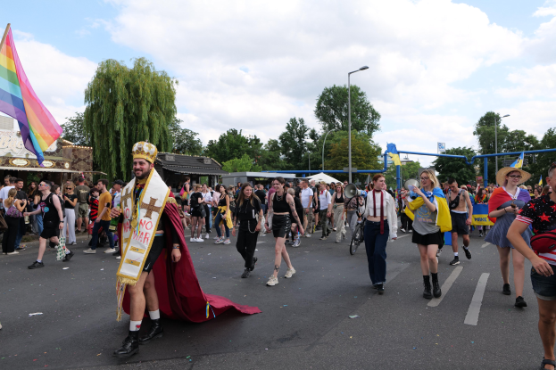 Eine Gruppe von Menschen marschiert bei der Gay Pride Parade 2018 mit einer Regenbogenflagge und Musikinstrumenten, einige tragen Mützen, vorbei an Laternenmasten, Bäumen, Schuppen und einem bewölkten Himmel.
