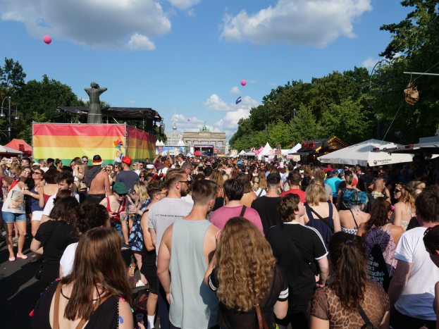 Eine große Menschenmenge, die eine Straße mit Zelten, Bäumen, Pfählen, Lichtern und Gebäuden entlanggeht, mit einem bewölkten Himmel und Ballons im Hintergrund beim Christopher Street Day in Berlin.