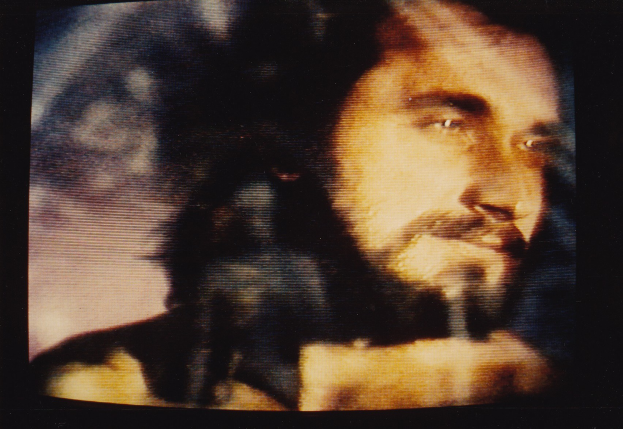 Ein Mann mit langem dunklem Bart und Schnurrbart, der ein weißes Hemd trägt, hat einen ernsten Gesichtsausdruck, während er direkt in die Kamera auf einem Fernseher mit dunklem Hintergrund schaut.