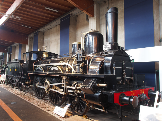 Alte Dampflokomotive in einem Museum ausgestellt mit umgebenden Schienenstrassen, Informationsschildern und anderen Ausstellungen, vor einer Wand mit Deckenleuchten.