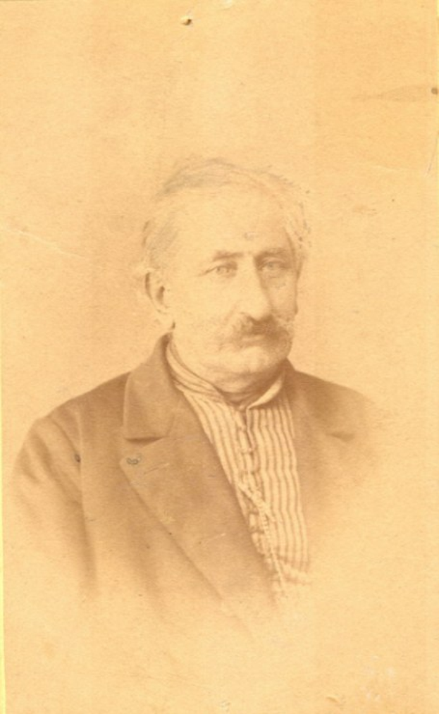 Ein Schwarz-Weiß-Porträt eines älteren Mannes mit Schnurrbart, der einen Mantel trägt und ein starkes Kinn, eine markante Nase, dunkle Augen und einen nachdenklichen Ausdruck mit leicht geöffneten Lippen hat.