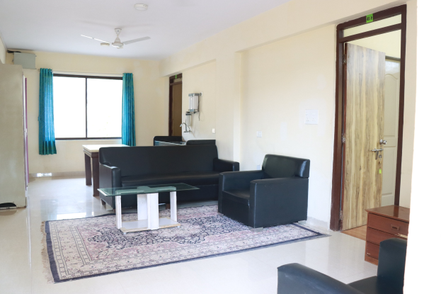 Ein Wohnzimmer mit einer Sitzgruppe, Beistelltischen, einem Teppich, einem wandmontierten Flachbildfernseher, Vorhängen, Deckenleuchten und einer Tür.