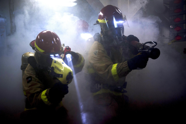 Feuerwehrleute in Schutzausrüstung spritzen Wasser auf eine rauchgefüllte Wand, während ein Schild mit Text links sichtbar ist.