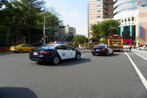 Ein Polizeiwagen fährt auf einer Straße mit hohen Gebäuden, Bäumen, Laternen, Ampeln und Menschen, unter einem bewölkten Himmel.