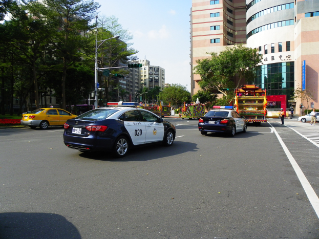 Ein Polizeiwagen fährt auf einer Straße mit hohen Gebäuden, Bäumen, Laternen, Ampeln und Menschen, unter einem bewölkten Himmel.