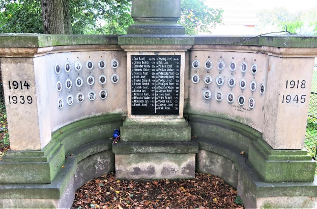 Ein Holocaust-Denkmal in einem jüdischen Friedhof in Berlin, mit einer Tafel mit Text und Zahlen, umgeben von Bäumen, einem Zaun und verstreuten trockenen Blättern.
