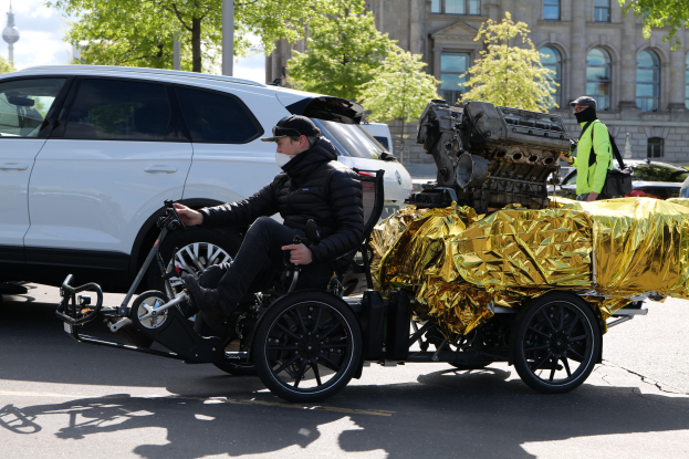 Ein Mann im Rollstuhl mit einem großen Motor auf dem Rücken, der ein Objekt hält, auf einer Straße umgeben von Fahrzeugen, Bäumen, Gebäuden, Polen und einem klaren blauen Himmel.