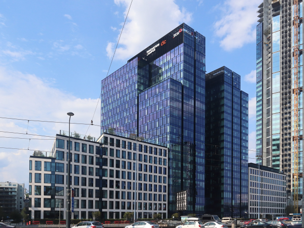 Zentrale der Deutschen Bank in Frankfurt, Deutschland, ein großes mehrstöckiges Gebäude mit zahlreichen Fenstern, umgeben von städtischer Infrastruktur einschließlich Straßenlaternen, Elektrofahrzeugen, Grünflächen und einem bewölkten Himmel.
