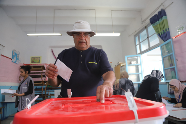 Ein Mann mit Hut stimmt bei einer Wahllokal ab, vor einer roten Wahlurne stehend, mit einem Zettel in der Hand, während andere an Tischen sitzen und schreiben, vor einem Fenster mit einem Vorhang.