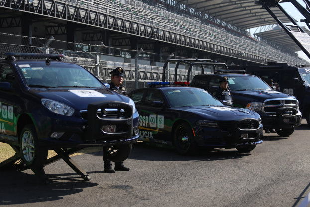 Eine Gruppe von Polizeiwagen vor einer Rennstrecke geparkt, uniformierte Beamte auf der Straße stehend und ein Stadion mit Geländern und Maschendrahtzaun im Hintergrund.