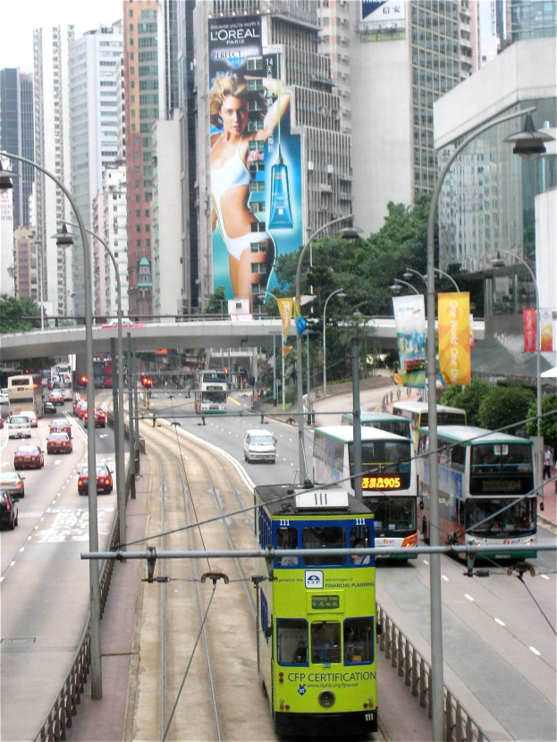 Eine belebte Stadtstraße mit Doppeldeckerbussen, Autos und anderen Fahrzeugen, die Laternen, Schilder, Plakate, Bäume, eine Brücke, hohe Gebäude und einen klaren blauen Himmel mit Text am unteren Rand zeigt.