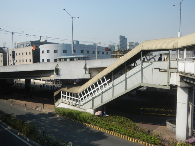 Fußgängerbrücke über einer Straße mit Menschen, umgeben von Gebäuden, Straßeninfrastruktur, Fahrzeugen, Grünflächen und einem klaren Himmel.