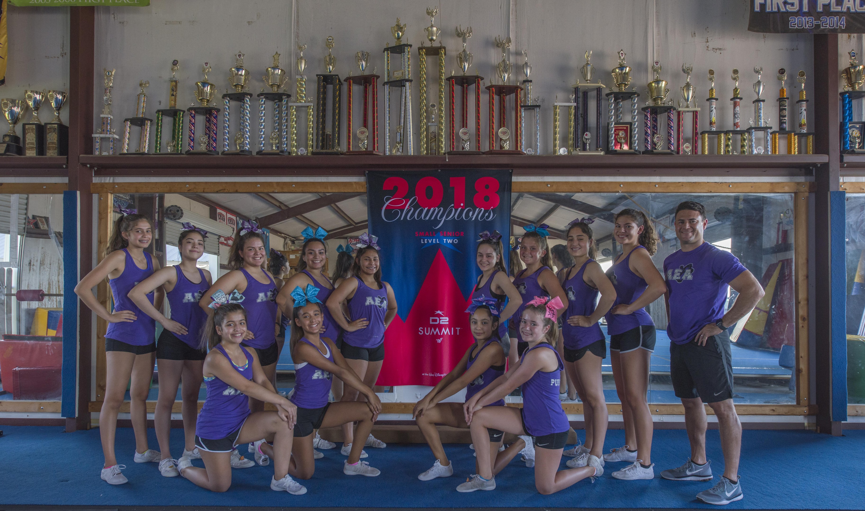 Gruppe von Cheerleadern in lila T-Shirts und schwarzen Shorts, die vor einer Vitrine mit Trophäen posen, im Hintergrund ein Banner mit der Aufschrift "2018 Summit Cheerleading Champions".